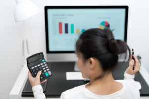 Woman with black hair sits at a computer looking at a calculator in her right hand