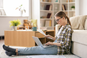 Feature image of a young white woman sitting on the floor, looking at a sheet of paper in her hand while typing on a laptop