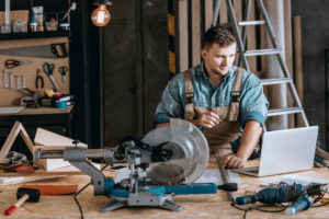 A white man with brown hair uses an electric saw to cut into a piece of wood